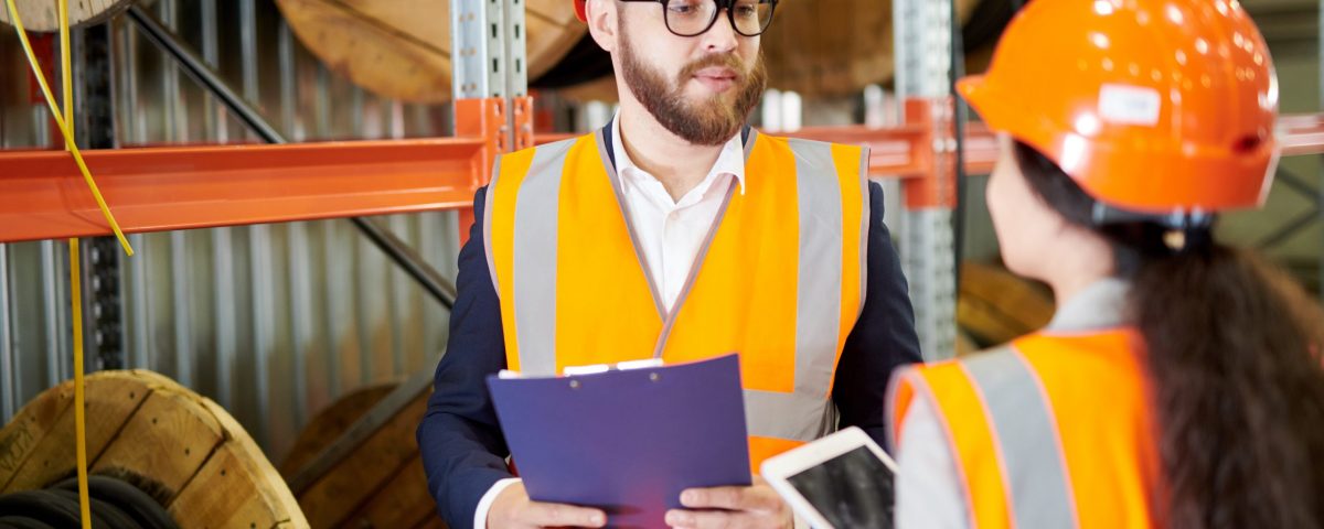 Portrait of modern bearded businessman wearing hardhat listening to factory employee while standing in workshop and holding clipboard, copy space