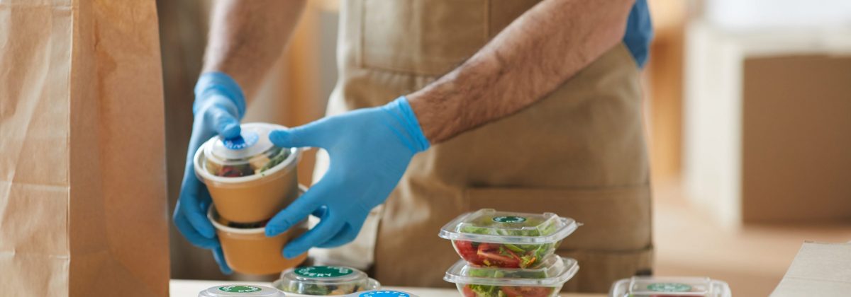 Closeup of unrecognizable worker wearing protective gloves packaging orders at wooden table in food delivery service, copy space